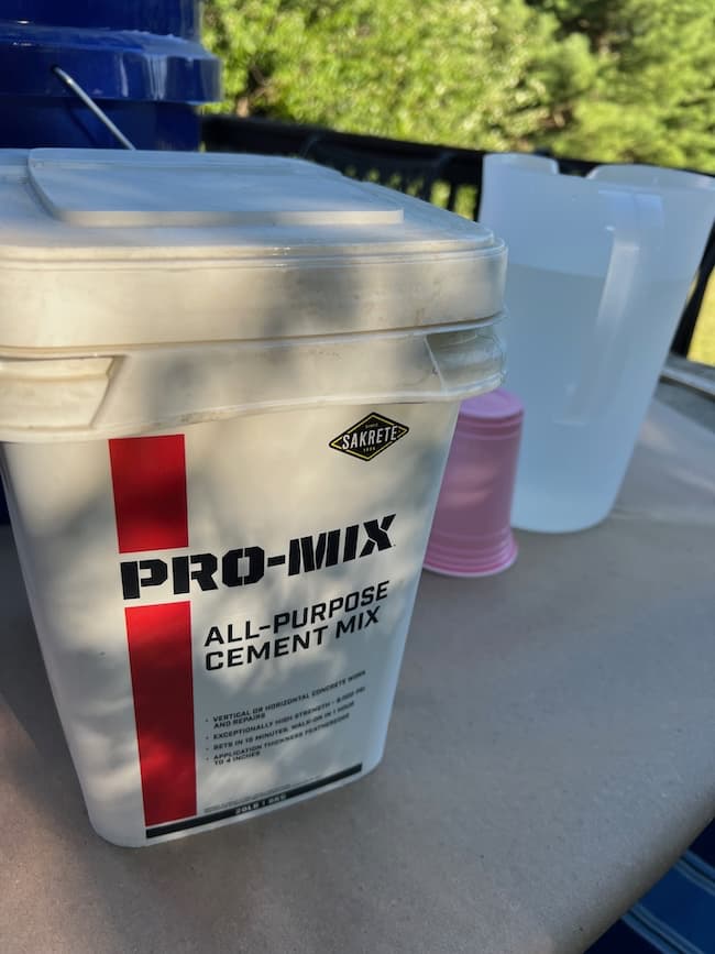 A container of Sakrete Pro-Mix All-Purpose Cement Mix is on a table outdoors, perfect for a DIY cement planter bowl project. Plastic cups and a pitcher of water are in the background, surrounded by greenery. The table is set in a bright, sunny environment.