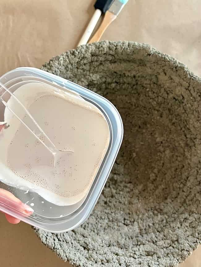 A hand holds a small square plastic clear container, full of off white paint that is stirred with a clear plastic spoon. The container is held over a rustic textured DIY cement planter that is gray in color.