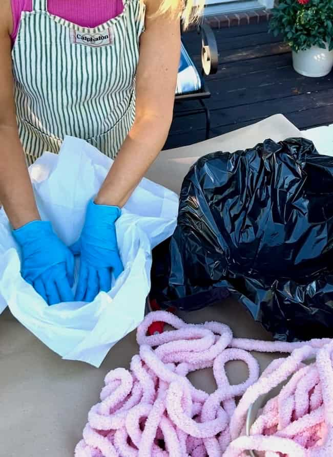 A person wearing blue gloves and a striped apron is arranging white tissue paper on a table alongside materials for a DIY cement planter bowl. Nearby, there is a black plastic bag and a pile of pink, fluffy rope.