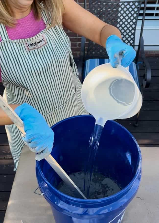 A person wearing blue gloves and a striped apron is pouring a white liquid from a bowl into a blue bucket, preparing to create a DIY cement planter bowl. They are holding a wooden stick in the bucket to mix the contents. The person is on a wooden deck, with a metal chair visible in the background.