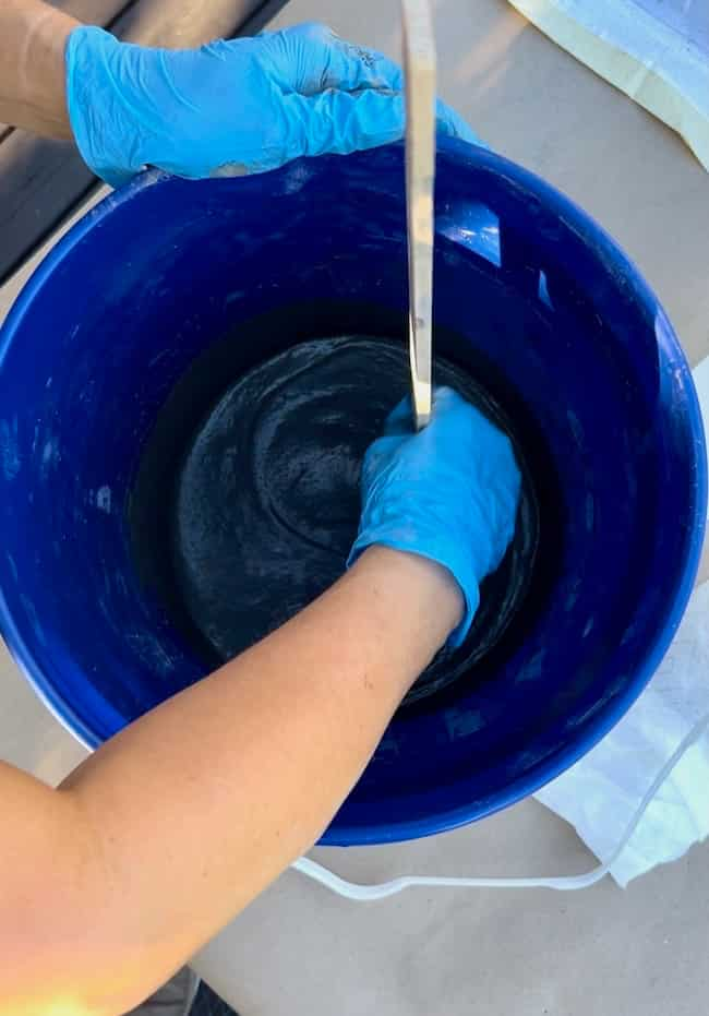 A person wearing blue gloves is mixing a thick, dark substance in a large blue bucket using a long, thin tool. The bucket is placed on a light-colored surface, surrounded by various items needed for their DIY cement planter bowl project.