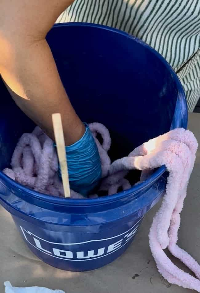 A person wearing a blue glove swirls a stick in a blue Lowe's bucket filled with a pink, fluffy material, possibly preparing it for a DIY cement planter bowl. The person's arm and part of their striped clothing are visible as they mix the contents inside the bucket.