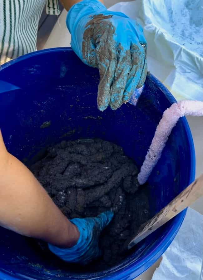 A person wearing blue gloves is mixing or handling dark, mud-like material in a blue bucket, likely creating a DIY cement planter bowl. The mixture appears thick and clumpy. A pink pipe cleaner is visible on the side of the bucket, and part of a wooden stick is partially shown on the right.