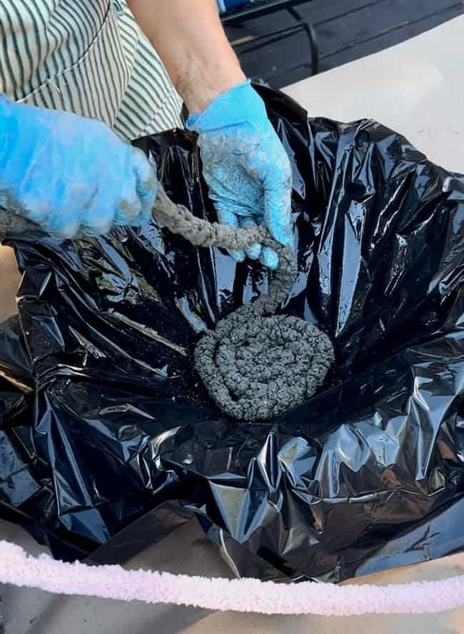 A person wearing blue gloves molds a coil of concrete inside a black plastic-lined container, carefully shaping it into a spiral pattern to create a DIY cement planter bowl. The background includes parts of their clothing and a surface draped in fabric.