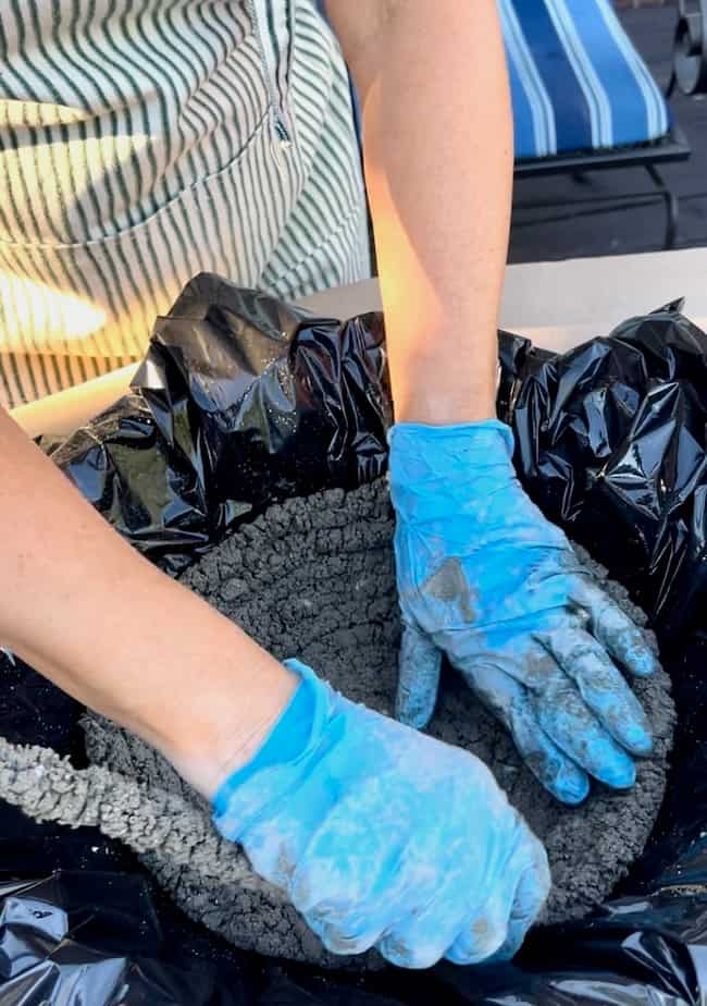 Person wearing blue gloves, pressing wet concrete mixture into a black plastic-lined container to create a DIY cement planter bowl. They are dressed in a striped shirt. The concrete appears fresh and malleable, with a patio chair visible in the background.