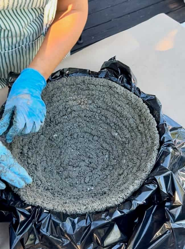 A person wearing a striped apron and blue gloves is molding a circular concrete structure. The structure, potentially a DIY cement planter bowl, is lined with a black plastic sheet and appears to be in the early stages of setting. The person is smoothing the inner surface of the concrete.