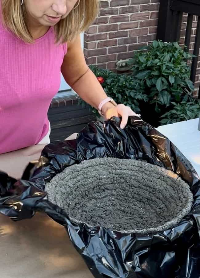 A person wearing a pink sleeveless top is holding a black plastic sheet covering a round, gray concrete form. They appear to be crafting a DIY cement planter bowl at a table with plants and a brick wall in the background.