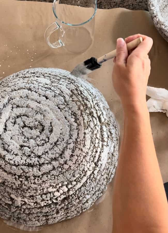 Top-down view of a person's hand painting a textured, dome-shaped object with a paintbrush. The DIY cement planter bowl, which looks like a concrete or plaster sculpture, is on a brown paper-covered surface. A clear glass mug is also visible in the background.