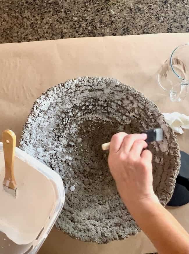 A person is painting the inside of a textured, round concrete object, creating a DIY cement planter bowl with a small paintbrush. Nearby are a larger paintbrush, a plastic container, and a clear measuring cup placed on a brown paper-covered surface.