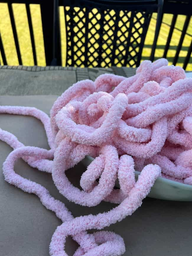 A large tangled bundle of soft, pink, fluffy yarn is placed in a white DIY cement planter bowl on a table. In the background, there is an outdoor setting with a black, patterned metal chair and a view of a grassy lawn.