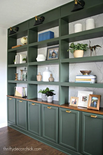 A bookcase painted in moody dark green, with closed cabinets at the bottom and open shelves on top, adorned with decorative items like plants, books, framed pictures, and vases. Brass hardware and three light fixtures add a touch of elegance. Text on image: thriftydecorchick.com.