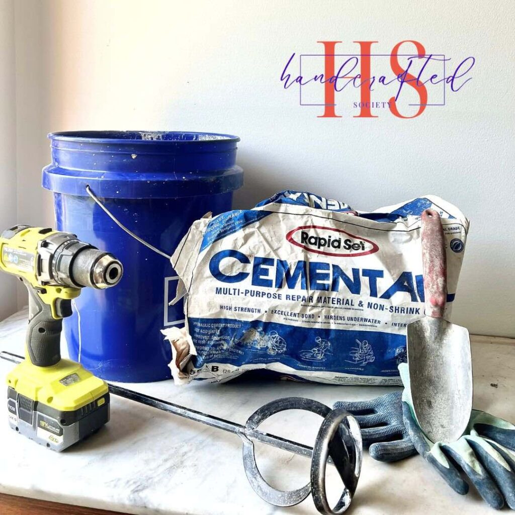 A blue bucket, a cement mix bag, a power drill, a trowel, and a mixing paddle are arranged on a white surface. A hose is coiled nearby. In the background, the words "IIS Handcrafted Society" are partially visible, hinting at an upcoming DIY cement planter bowl project.