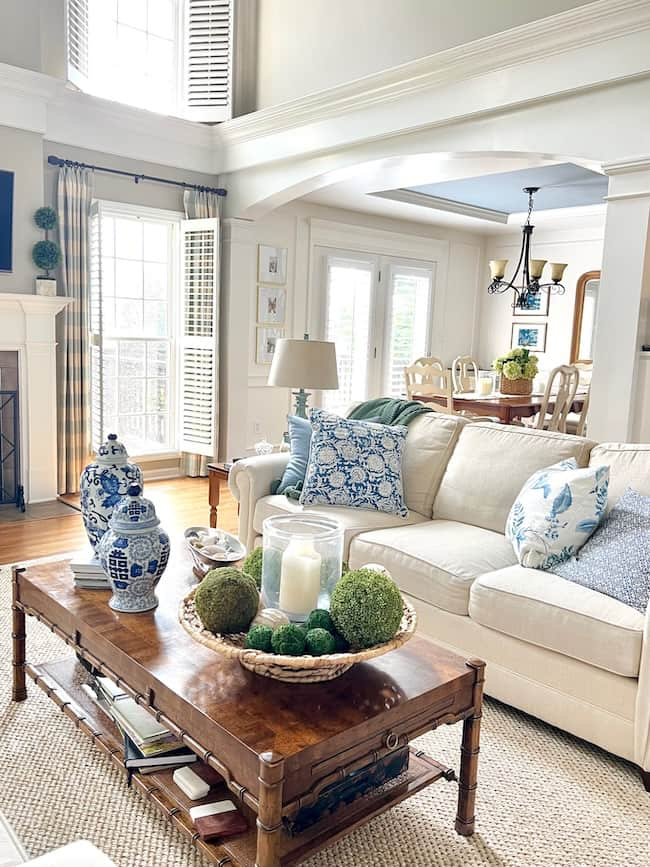 A spacious living room with high ceilings and large windows, featuring a white sofa adorned with blue and white throw pillows. The wooden coffee table holds decorative items, including candles and green moss balls. Moody paint colors enhance the ambiance. A dining area is visible in the background.