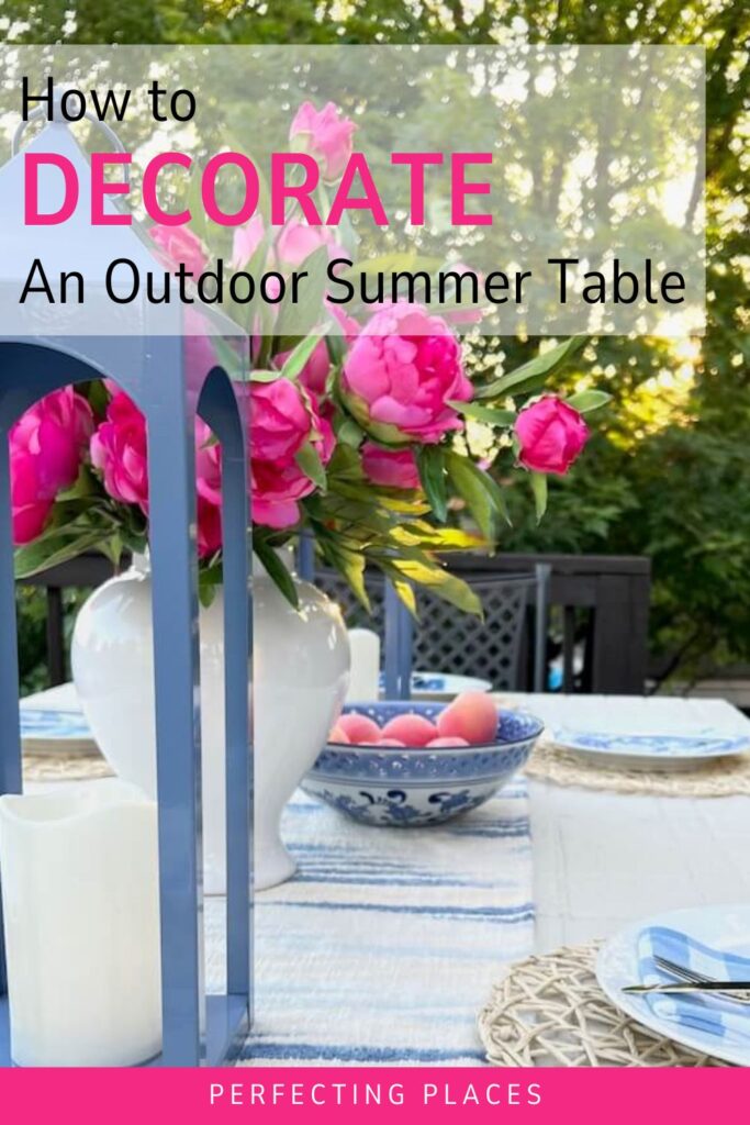 A beautifully decorated outdoor summer table set in a garden with a white vase holding vibrant pink flowers as the centerpiece, blue lanterns, and a bowl of peaches on a blue and white striped table runner. The text overlay reads "How to Set a Pretty Summer Table.