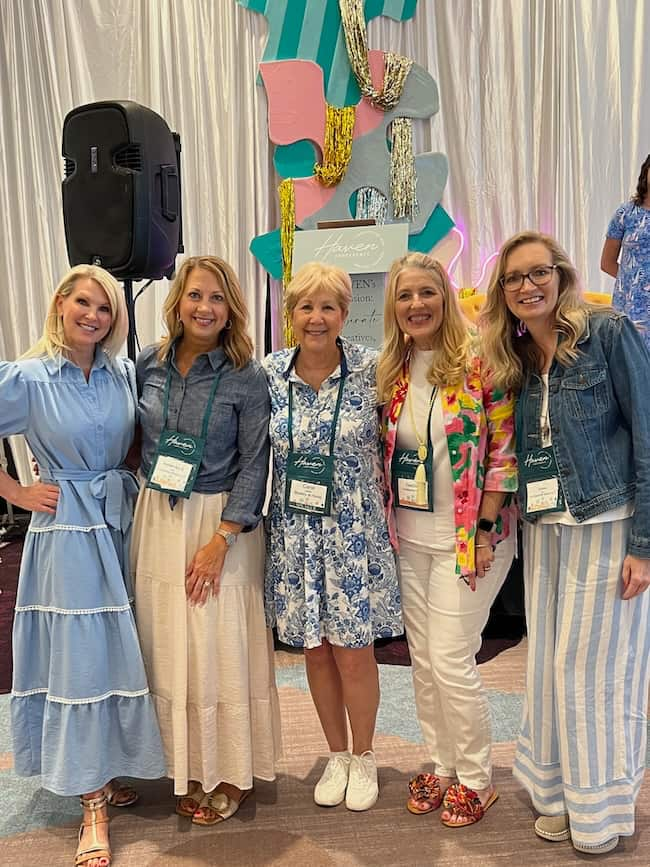 Seven women are standing together indoors, smiling at the camera. They are wearing a variety of colorful and patterned outfits, some with name tags around their necks. Behind them is a brightly decorated backdrop featuring abstract shapes and a large "7/20/24" in bold letters.