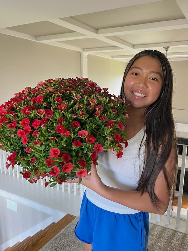 A woman with long dark hair, wearing a white tank top and blue shorts, smiles while holding a large, blooming plant with numerous small red flowers. She is standing indoors next to a white railing with a beige wall and ceiling in the background on what appears to be Seven on Saturday.