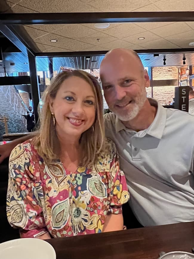 A smiling couple sits together in a restaurant called "Seven on Saturday." The woman has shoulder-length blond hair and is wearing a colorful, floral-patterned blouse. The man, who has a bald head and a short beard, is wearing a light gray polo shirt. The restaurant has a warm, inviting ambiance.