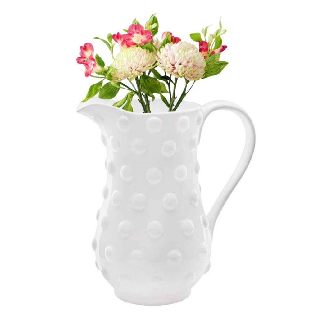 A white ceramic pitcher with a raised dot pattern holds a bouquet of pink and white flowers with green leaves. The pitcher has a curved handle and a spout. The flowers, arranged to appear fresh and vibrant, give a lively touch to the elegant, textured pitcher—the perfect centerpiece to set a pretty summer table.