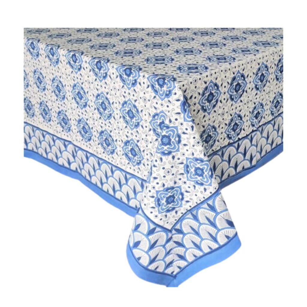 A rectangular table set a pretty summer table, covered with a blue and white patterned tablecloth featuring intricate floral and geometric designs. The tablecloth has a blue border with fan-like motifs along the edges.