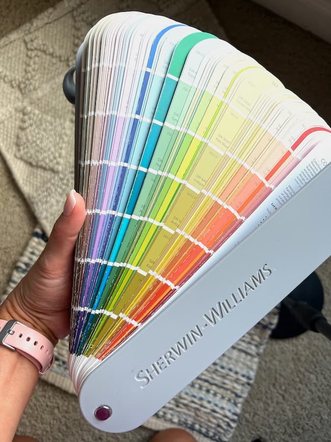 A hand holding a Sherwin-Williams paint color fan deck, displaying a wide array of moody paint colors arranged in a gradient from light to dark shades. The fan deck is spread out, revealing various hues including blues, greens, yellows, and reds.