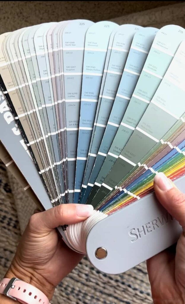 A person is holding a large Sherwin-Williams paint color fan deck, displaying various shades of moody blues, greens, and grays. The fan is fanned out showing multiple color swatches. The person's fingernails are painted pink, and they are wearing a pink watch.