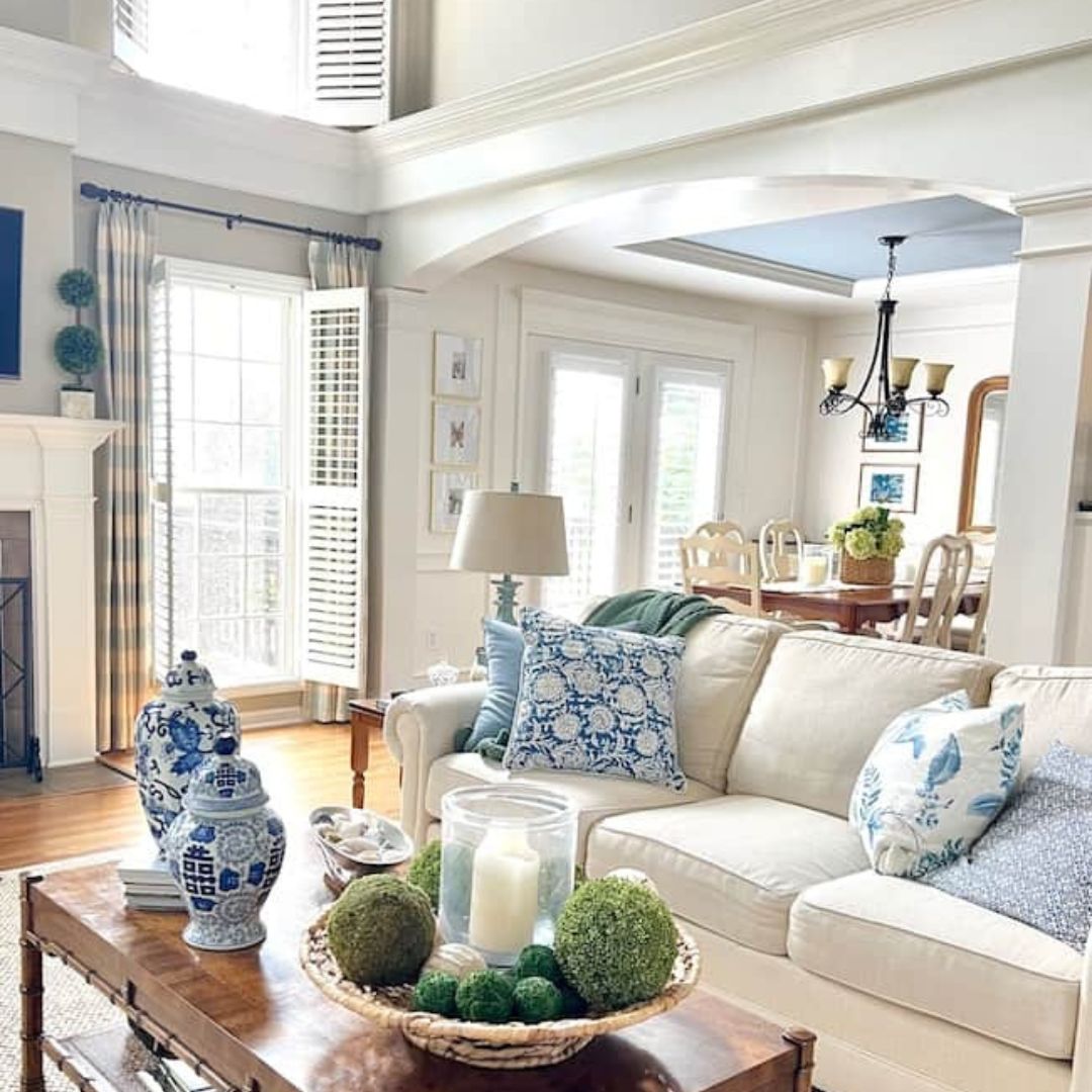 A bright and airy living room featuring a white sofa adorned with blue patterned pillows, beautifully contrasting moody paint colors. A wooden coffee table holds candles and decorative moss spheres. The space opens to a dining area with a chandelier and large windows, letting in natural light.