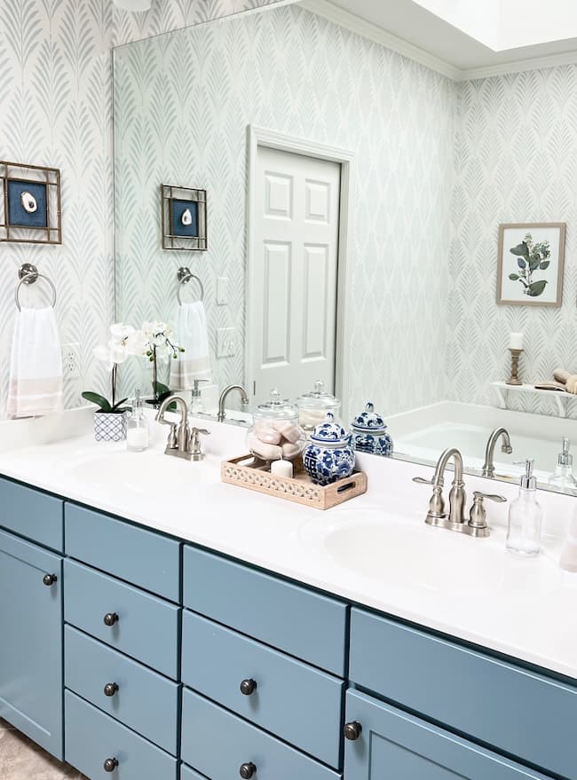 A bathroom with a large rectangular mirror reflecting a double sink vanity with blue cabinets painted in moody colors. The countertop features decorative items, including potted orchids, blue and white ceramic jars, and a tray with soaps and a rolled towel. The walls have light patterned wallpaper.