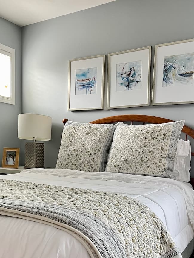 A cozy bedroom features a neatly made bed with a white and floral patterned quilt and matching pillows. Above the wooden headboard, three framed abstract art pieces add charm. A bedside table holds a beige lamp and a framed photo, set against light gray walls that contrast beautifully with moody paint colors elsewhere in the room.