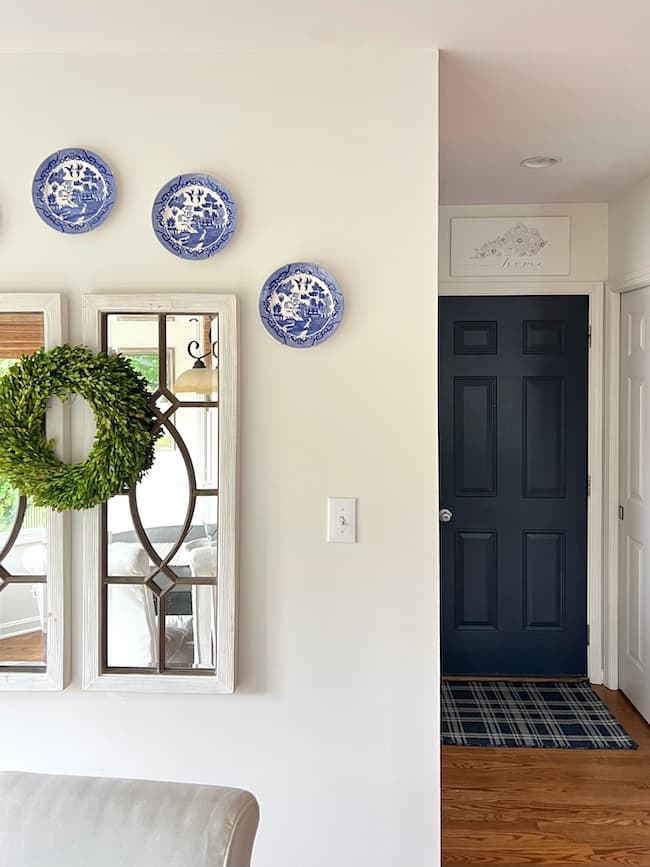 A cozy interior features a white wall with three blue decorative plates and two framed mirrors, one with a green wreath. The adjacent hallway showcases moody paint colors, including a dark blue front door with a small rug in front of it, set against warm wooden flooring.