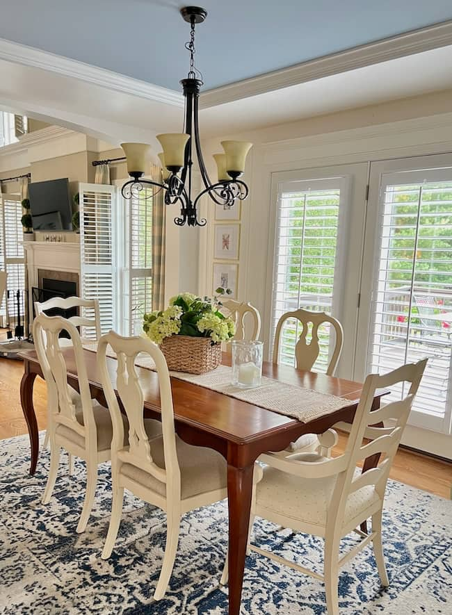 A dining room featuring a wooden table surrounded by six white chairs. A basket with green and white flowers serves as a centerpiece. The room, brightened by natural light from glass doors with white shutters, is beautifully contrasted by moody paint colors. A chandelier hangs overhead, and a patterned blue rug covers the floor.