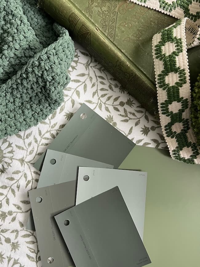 A cozy scene featuring moody paint colors in various shades of green swatches laid out on a white surface with a leafy pattern. A textured green blanket, a patterned green fabric, and an intricately designed green book are placed around the swatches.