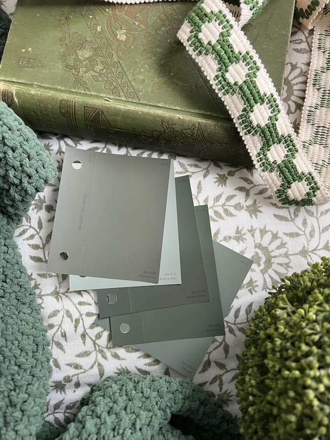 A collection of moody paint colors in green and neutral tones displayed on a floral-patterned surface, surrounded by textured green fabric, a green book with an ornate cover, and green and white patterned ribbons.