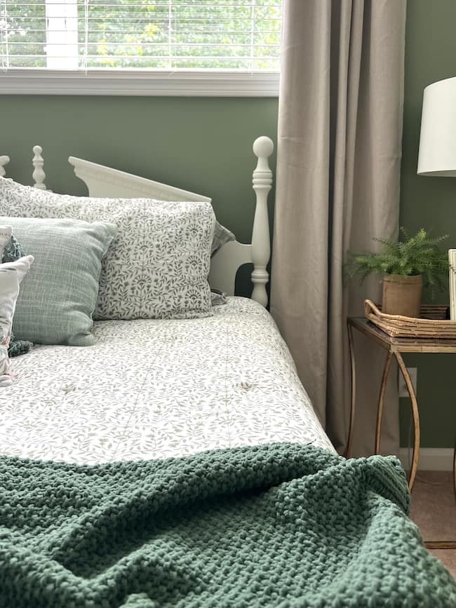 A cozy bedroom with a white bed featuring patterned bedding, green pillows, and a textured green throw blanket. A bedside table holds a small lamp and potted plant. Behind the bed is a window with blinds and curtains, framed by soft green walls accented with moody paint colors.