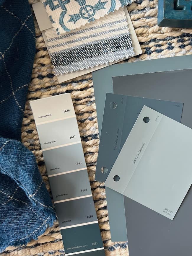 A flat lay image featuring a selection of blue-toned paint swatches, including moody paint colors, fabric samples with blue and white patterns, and a textured woven mat. The paint swatches display different shades from light to dark blue, arranged neatly alongside the fabrics.