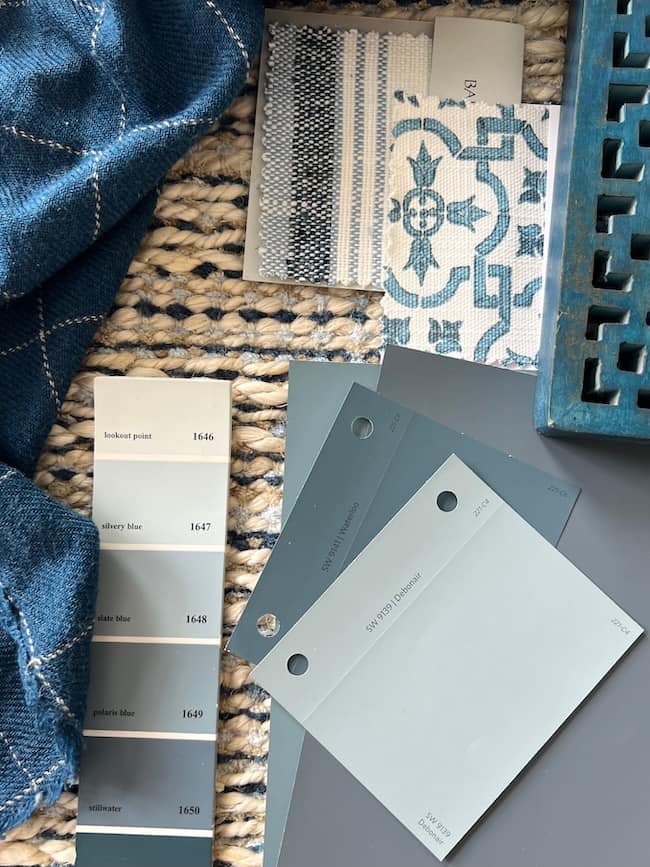 A photo showing various shades of moody blue and gray paint swatches, patterned fabric samples, and a textured rug. The colors range from light to dark blues and grays. There is also a portion of a decorative blue object in the corner.