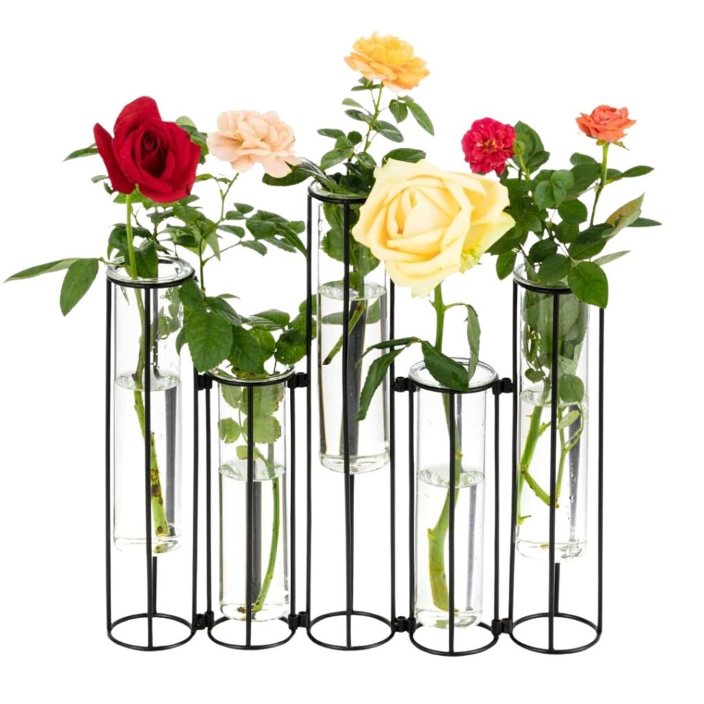 A black metal test tube vase holder contains six glass test tubes of varying heights, each holding a different colored rose, including red, pink, yellow, and peach. The roses are in bloom and surrounded by green leaves—a perfect centerpiece to set a pretty summer table.