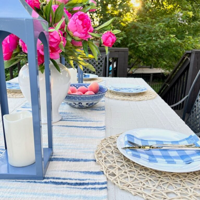 A sunlit outdoor dining table is set with patterned blue and white plates on woven placemats, silverware, a decorative vase with vibrant pink flowers, a bowl of peaches, and a white candle. The setting is adorned with a blue and white striped table runner. Trees in the background add a natural touch—perfect to set a pretty summer table.