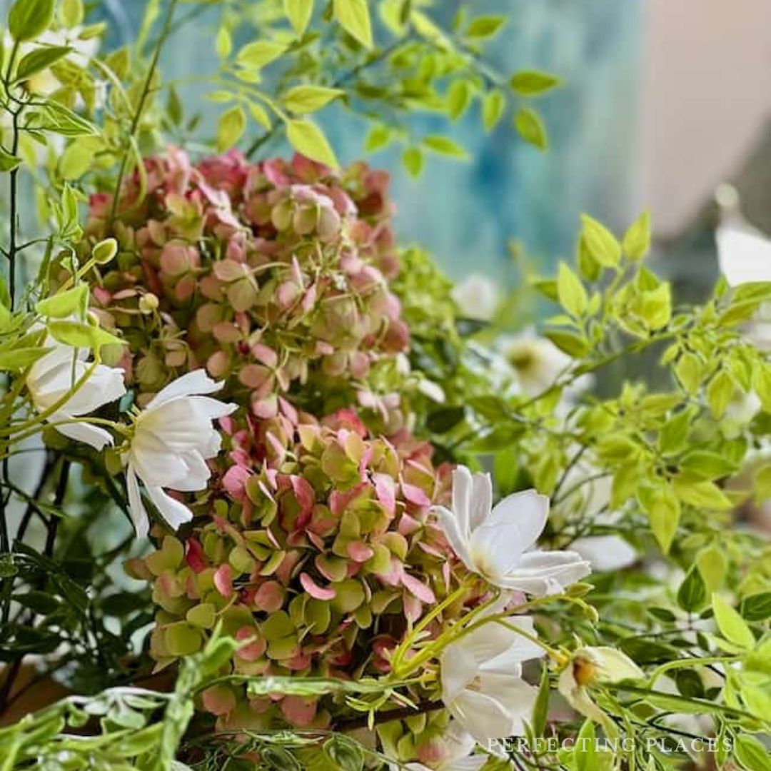 A close-up of a lush bouquet featuring hydrangeas with green and pink petals, interspersed with white daisies and surrounded by green foliage. The background, softly blurred with shades of blue and green, captures the essence of Seven on Saturday.