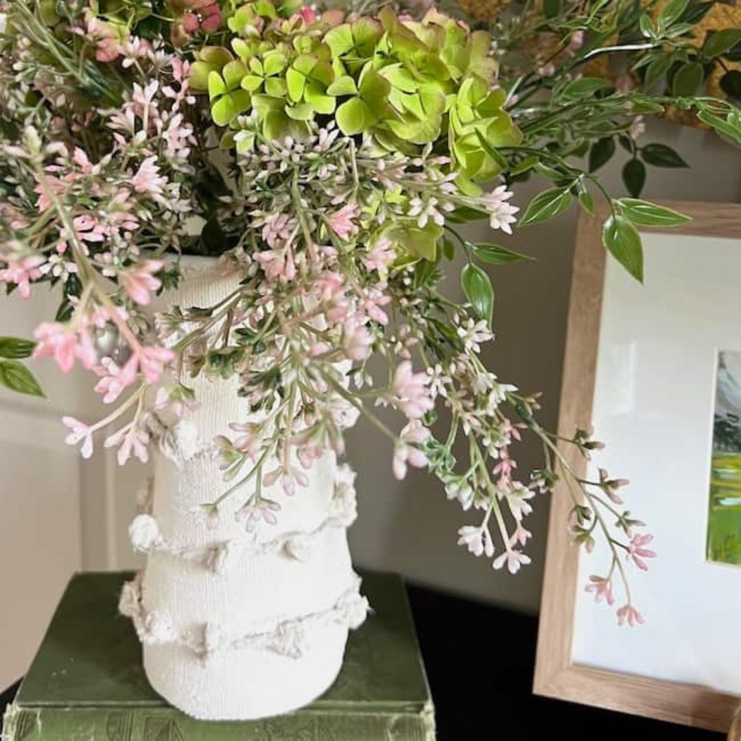 A decorative white vase, inspired by Anthropologie and wrapped in jute twine, is filled with lush greenery and delicate pink, green, and white flowers. It sits on a stack of books. The vase has a textured, rustic design. To the right, there is a framed picture partially visible.