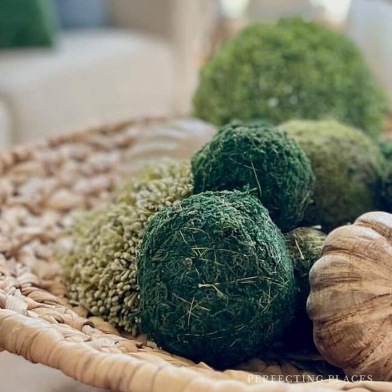 A woven basket containing assorted decorative balls of varying textures and green hues, including faux moss and dried plant materials, sits elegantly as part of home decor. The arrangement, perfect for a cozy Saturday, is placed on a blurred neutral background.