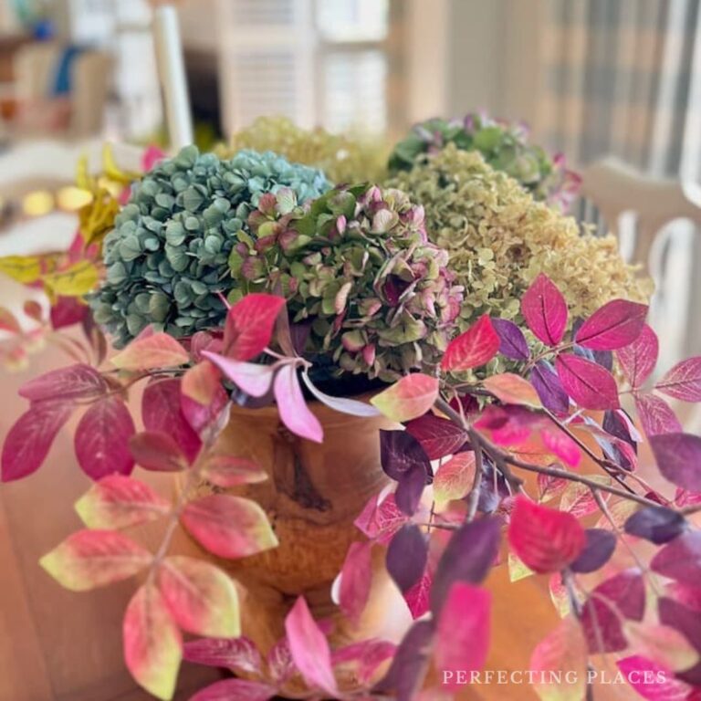 A vibrant autumn floral arrangement featuring green hydrangeas and pale brown flowers, surrounded by branches with bright pink and purple leaves, reminiscent of 'Seven on Saturday,' is displayed in a ceramic vase on a wooden table. Mark your calendars for 8/31/24 to experience this beauty.