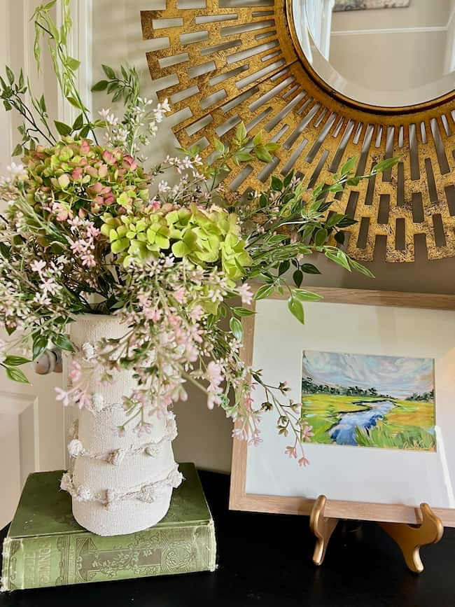 An interior scene shows a vase with a bouquet of flowers, set on top of a stack of books. Nearby, a framed landscape painting sits on a wooden stand. A large circular mirror with a sunburst pattern hangs on the wall behind the display, reflecting part of the room.