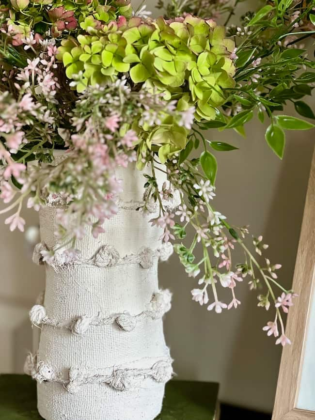 A close-up of a decorative vase wrapped in a textured white fabric with a knotted design, filled with a lush arrangement of green hydrangeas, pink flowers, and assorted greenery, set against a neutral indoor background.