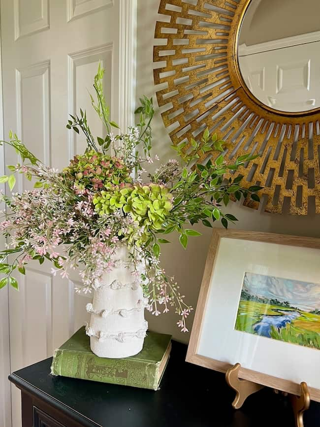 A floral arrangement featuring green and pink flowers is displayed in an Anthropologie inspired jute twine vase on a table. The table also holds a framed painting of a landscape and a green book. A circular gold mirror hangs on the wall above the table.