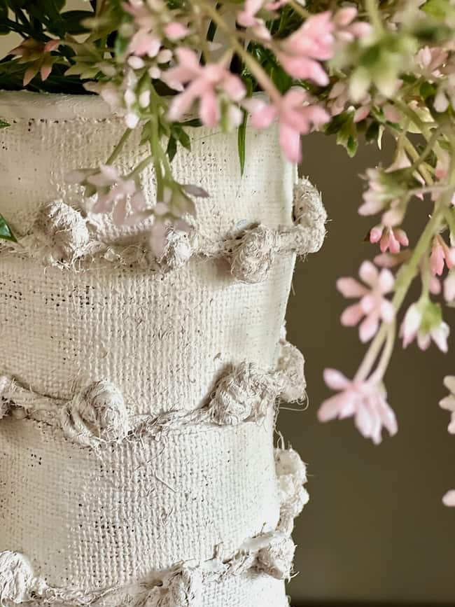 A close-up of a textured white vase decorated with raised, knotted details. The vase holds delicate pink flowers with green foliage, which cascade over the rim, creating a soft, natural look. The focus is on the vase's texture and the nearby flowers.