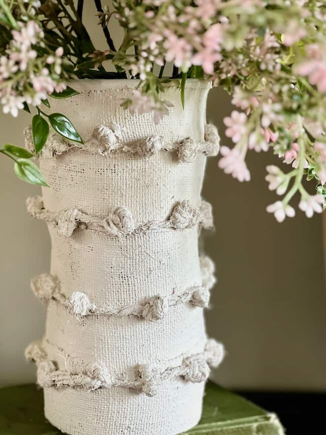 A white textured vase adorned with small pom-poms arranged in horizontal lines holds a bouquet of pink and white flowers with green leaves. The vase is set against a neutral background.