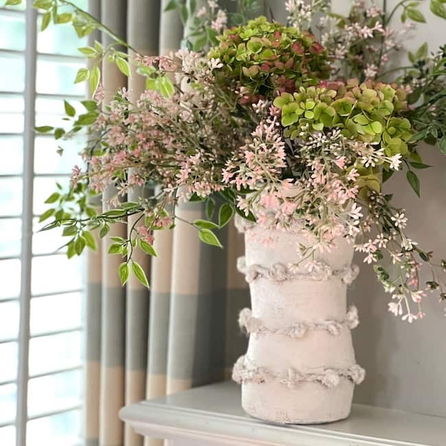 A white ceramic vase wrapped with Anthropologie-inspired jute twine holds a bouquet of green and pink hydrangeas, and delicate pink flowers with greenery. The vase is placed on a white surface, and the background features a window with striped curtains.