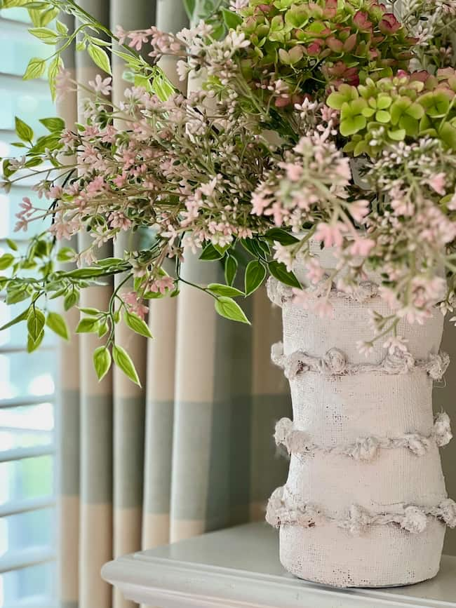 A decorative, Anthropologie-inspired white vase embellished with textured details and wrapped in jute twine holds an arrangement of green and pink hydrangeas, interspersed with small pink flowers and green leaves. The vase is placed on a light-colored surface against a backdrop of striped curtains and a sunlit window.