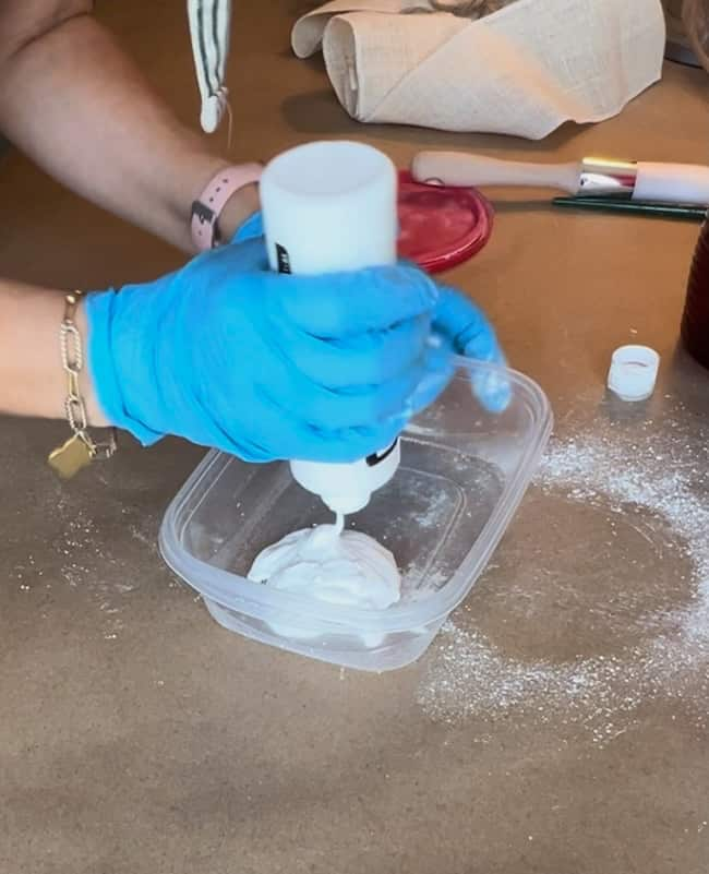 A person wearing blue gloves is squeezing a bottle of white substance into a clear plastic container on a table scattered with powder. Various items, including an Anthropologie inspired jute twine vase, a red lid, and kitchen tools, can be seen in the background.