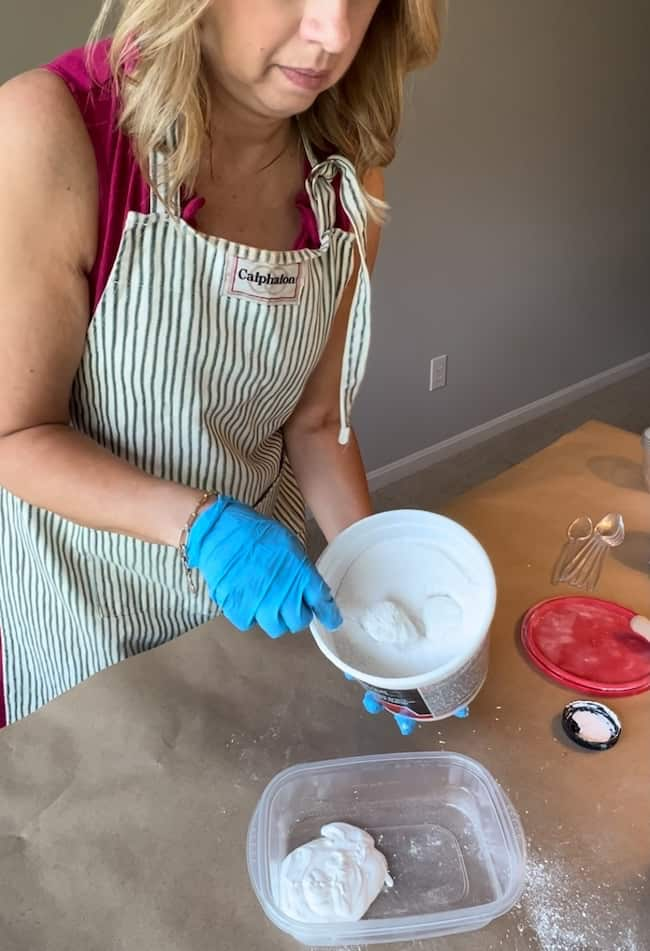 A person wearing a striped apron and blue gloves is scooping white plaster from a bucket into a clear plastic container. On the table in front of them are an Anthropologie inspired jute twine vase, a red lid, a whisk, and some scattered plaster.